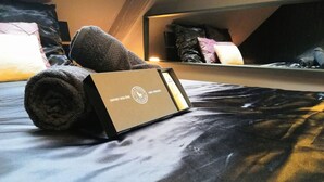 Een net opgemaakt bed met een opgerolde handdoek en een doos met de tekst 'COFFRET BIEN-ÊTRE' in een kamer met een schuine balkenkap en een spiegel die het bed en de kussens reflecteert.