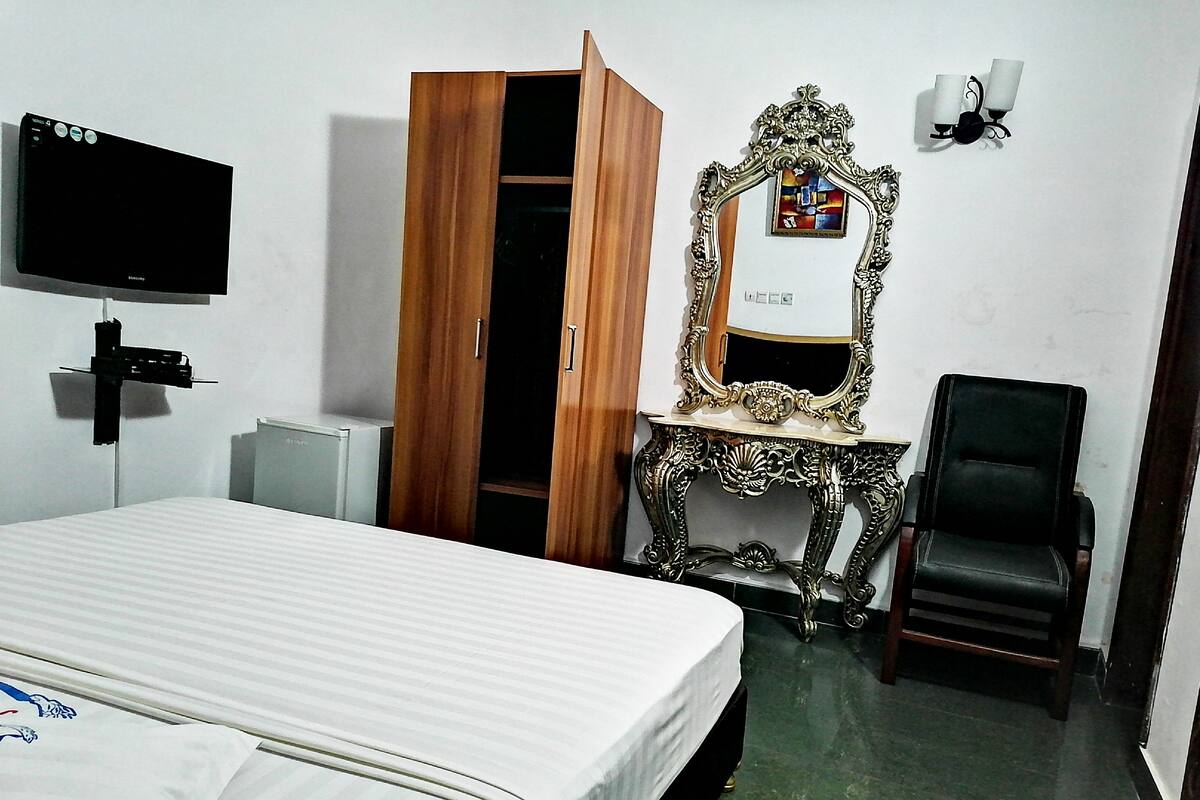 A hotel room with a bed, a wooden wardrobe, a vanity with an ornate mirror, a black chair, and a flat-screen TV mounted on the wall.