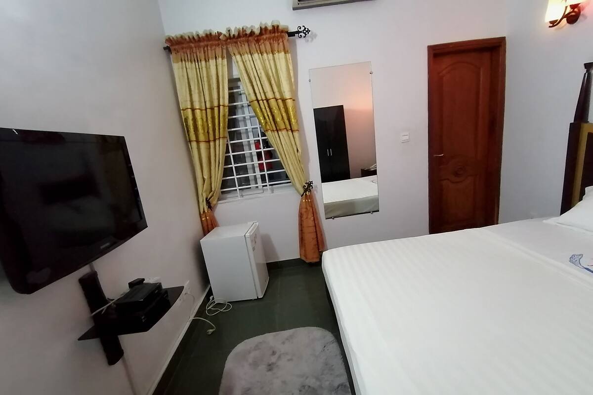 A hotel room with a bed, a television, a small refrigerator, a window with curtains, a mirror, and a door.