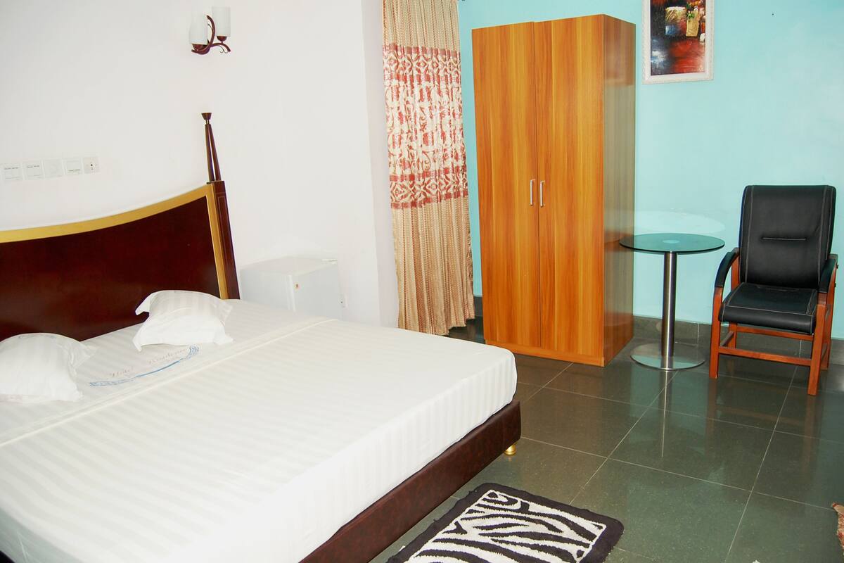 A bedroom with a bed, a wooden wardrobe, a small round table, and a black chair.