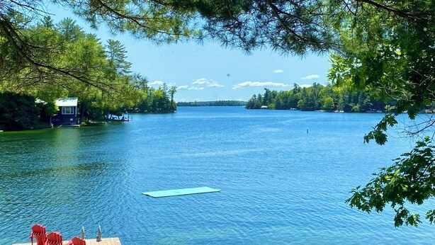A wooden dock with red chairs and a table, a clear blue lake, and a small island with a building. - A Cottage on a Private Island Big Rideau Lake (Perth)