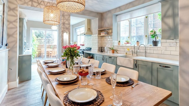 Una cocina moderna con comedor, que presenta una larga mesa de madera preparada para una comida, iluminación colgante y vistas al exterior.