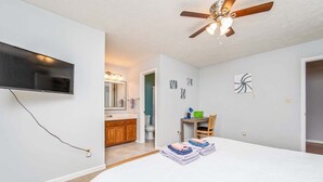 Un dormitorio con cama, ventilador de techo, televisor montado en la pared y un pequeño escritorio con silla.