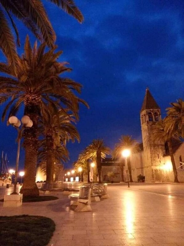A nighttime view of a square with palm trees, a historic building with a tower, and street lamps.