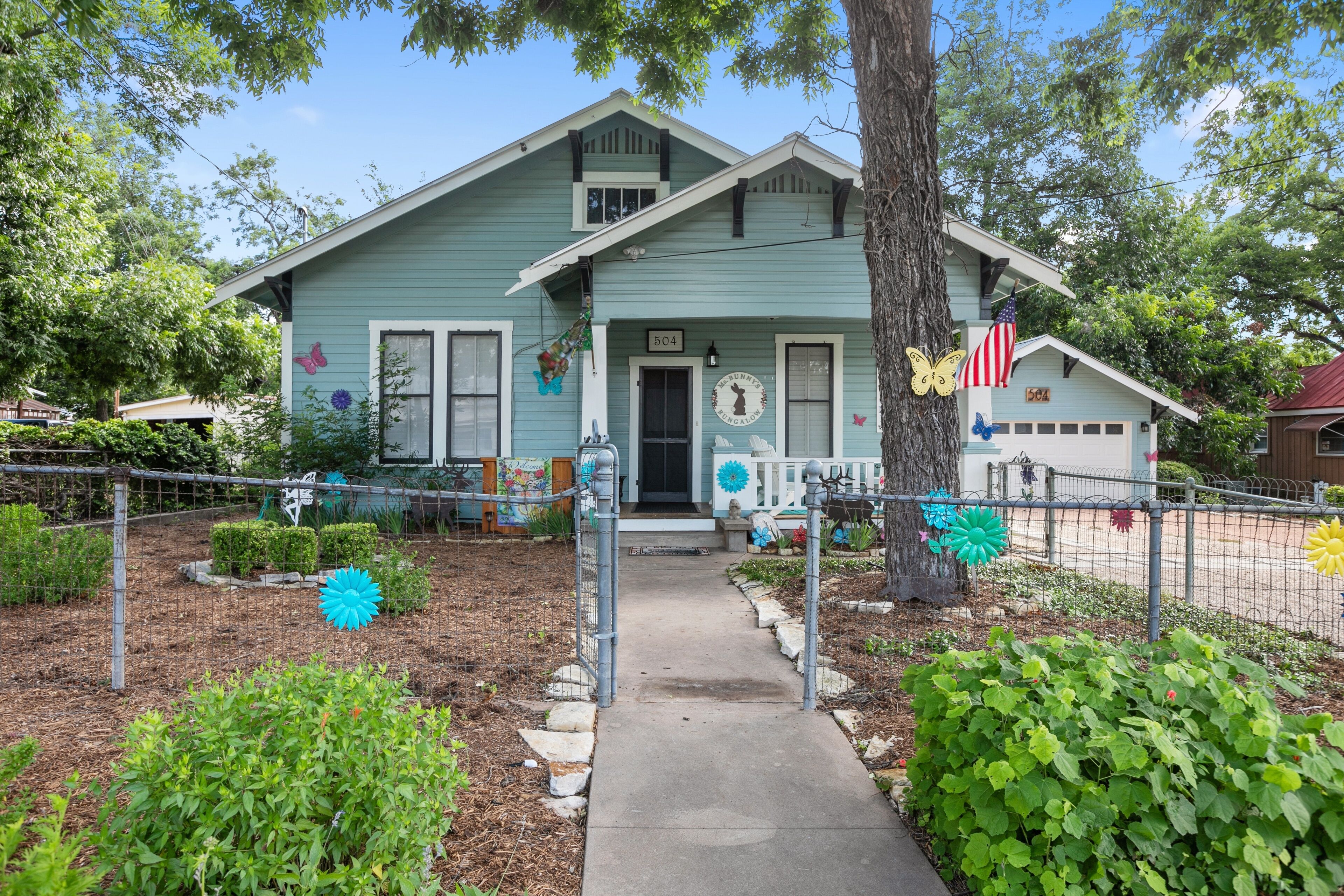 A light blue house with a front porch, a mailbox, and a small garden with decorative flowers.