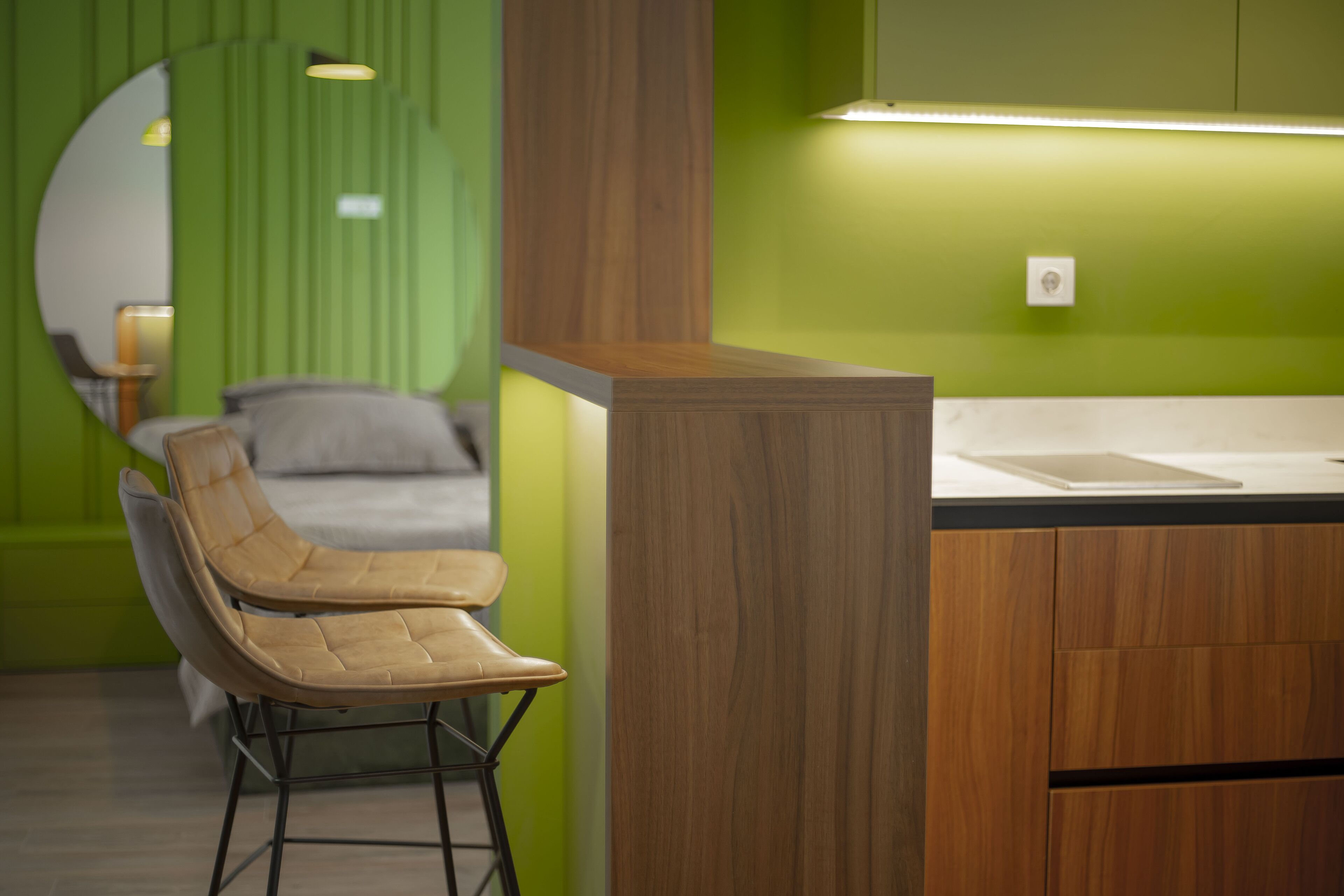 A modern room with a wooden reception desk, a green accent wall, a bed with a grey headboard, and a chair.