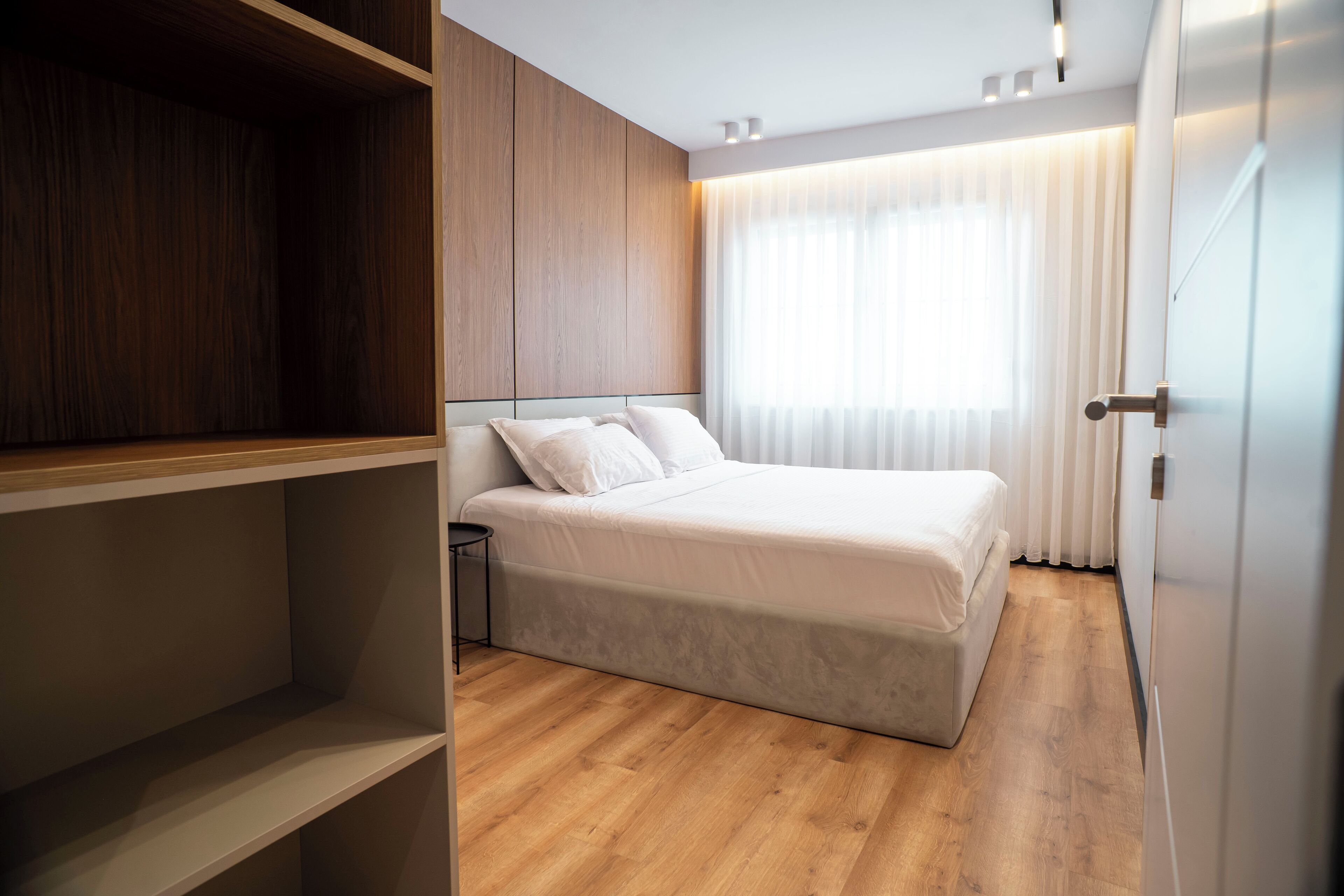 Un dormitorio moderno con un armario de madera, una cama con ropa de cama blanca y un gran ventanal con cortinas de gasa.