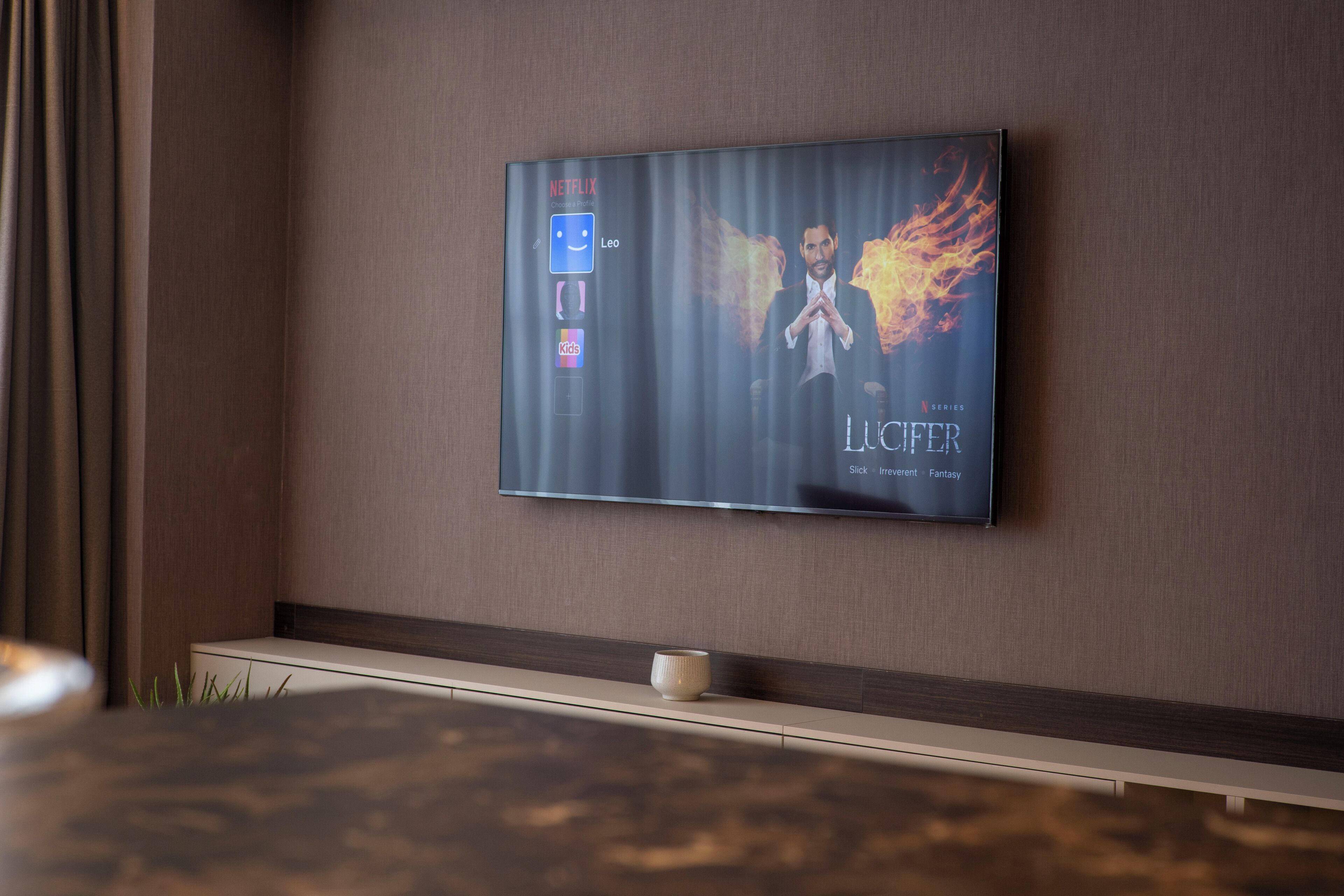Un televisor de pantalla plana montado en la pared, con la interfaz de Netflix mostrando la serie "Lucifer" y una sección de "Niños".