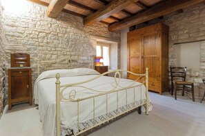 A bedroom with a stone wall, a wooden bed frame, a wooden wardrobe, and a chair.
