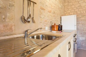 Una cucina con un lavandino in acciaio inox, utensili pensili, un frigorifero bianco e un muro in pietra.