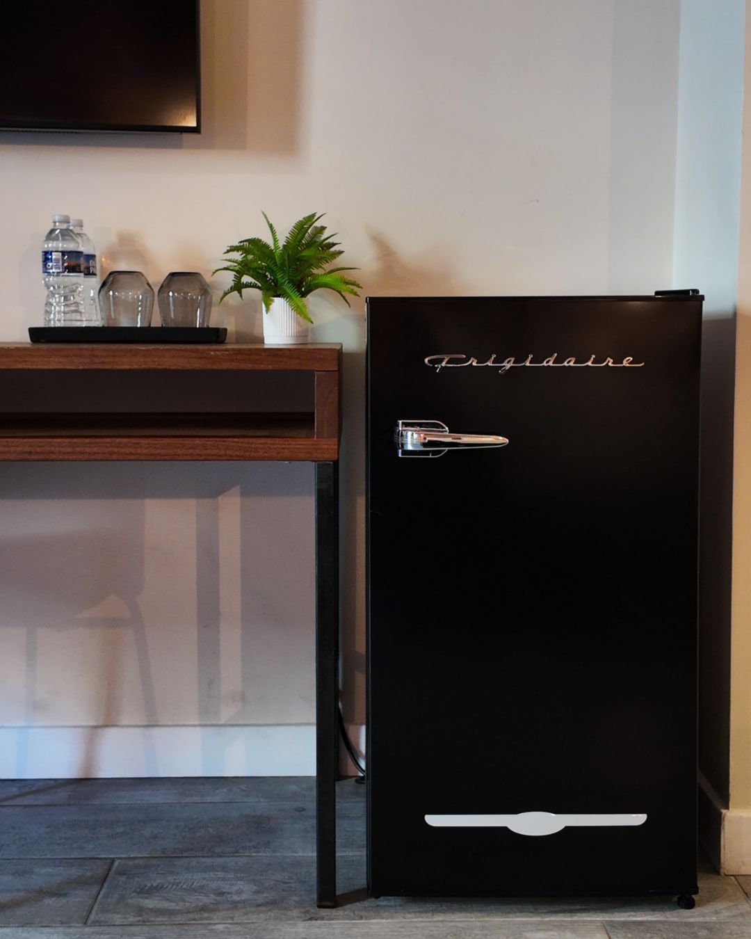 Ein schwarzer Kühlschrank mit dem Schriftzug 'Frigidaire' auf der Tür, neben einem Holztisch mit einem Topfpflanzen und zwei Glasflaschen.