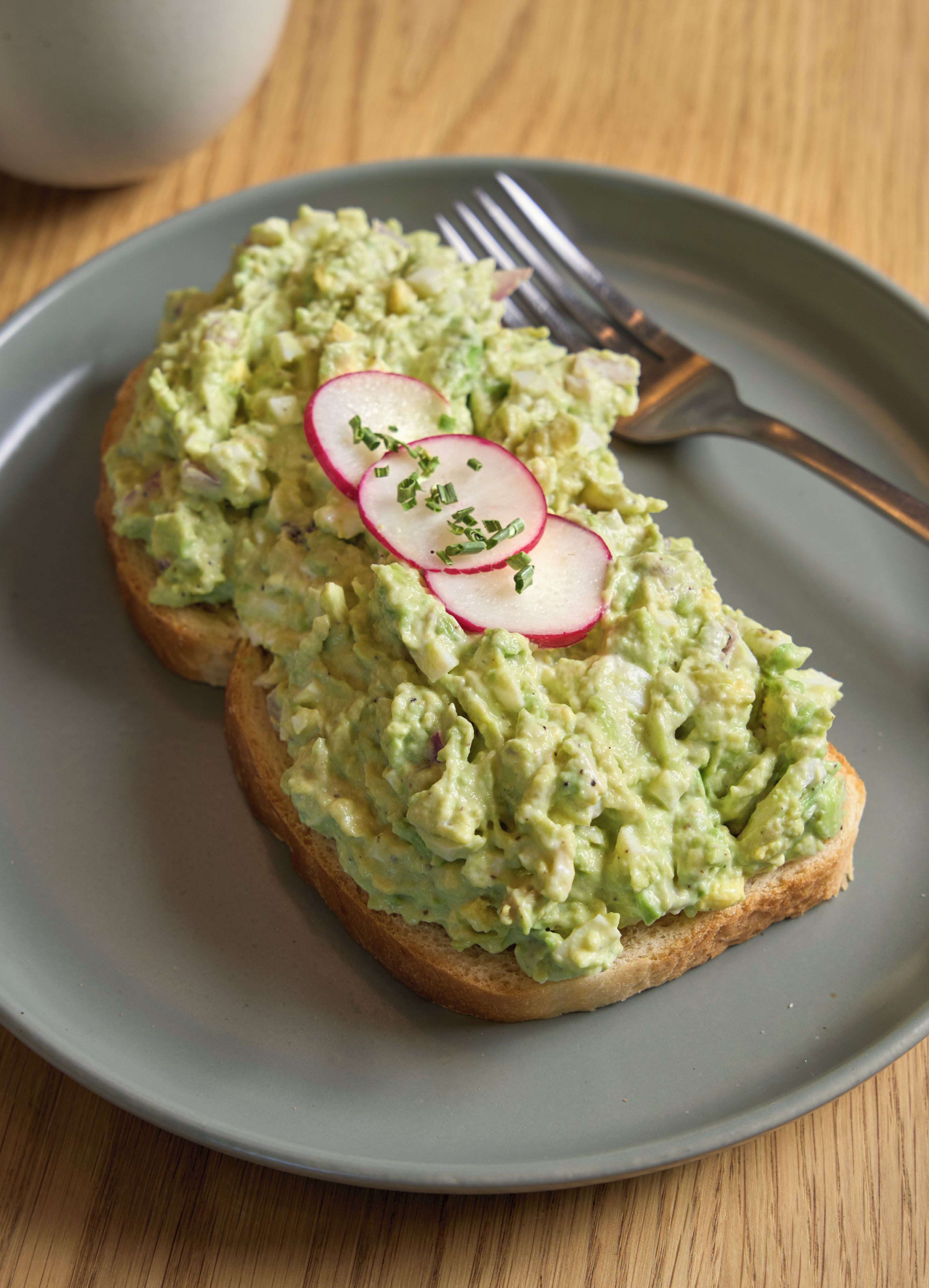 Ein Teller mit zwei Toastscheiben, belegt mit Avocadoaufstrich und Radieschenscheiben, dazu eine Gabel auf einem Holztisch.