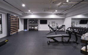 A gym with exercise equipment, a weight rack, and a wall-mounted TV. - The Maj Hotel (Philadelphia)
