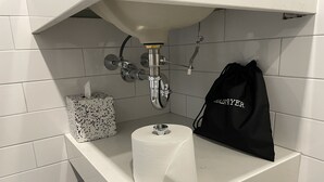 A bathroom sink with a paper towel holder, a toilet paper roll, and a black bag hanging on the wall. - The Maj Hotel (Philadelphia)
