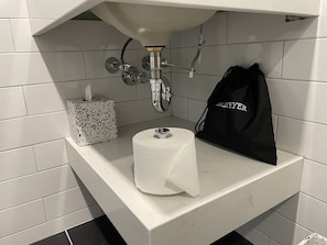A bathroom sink with a paper towel holder, a toilet paper roll, and a black bag hanging on the wall. - The Maj Hotel (Philadelphia)
