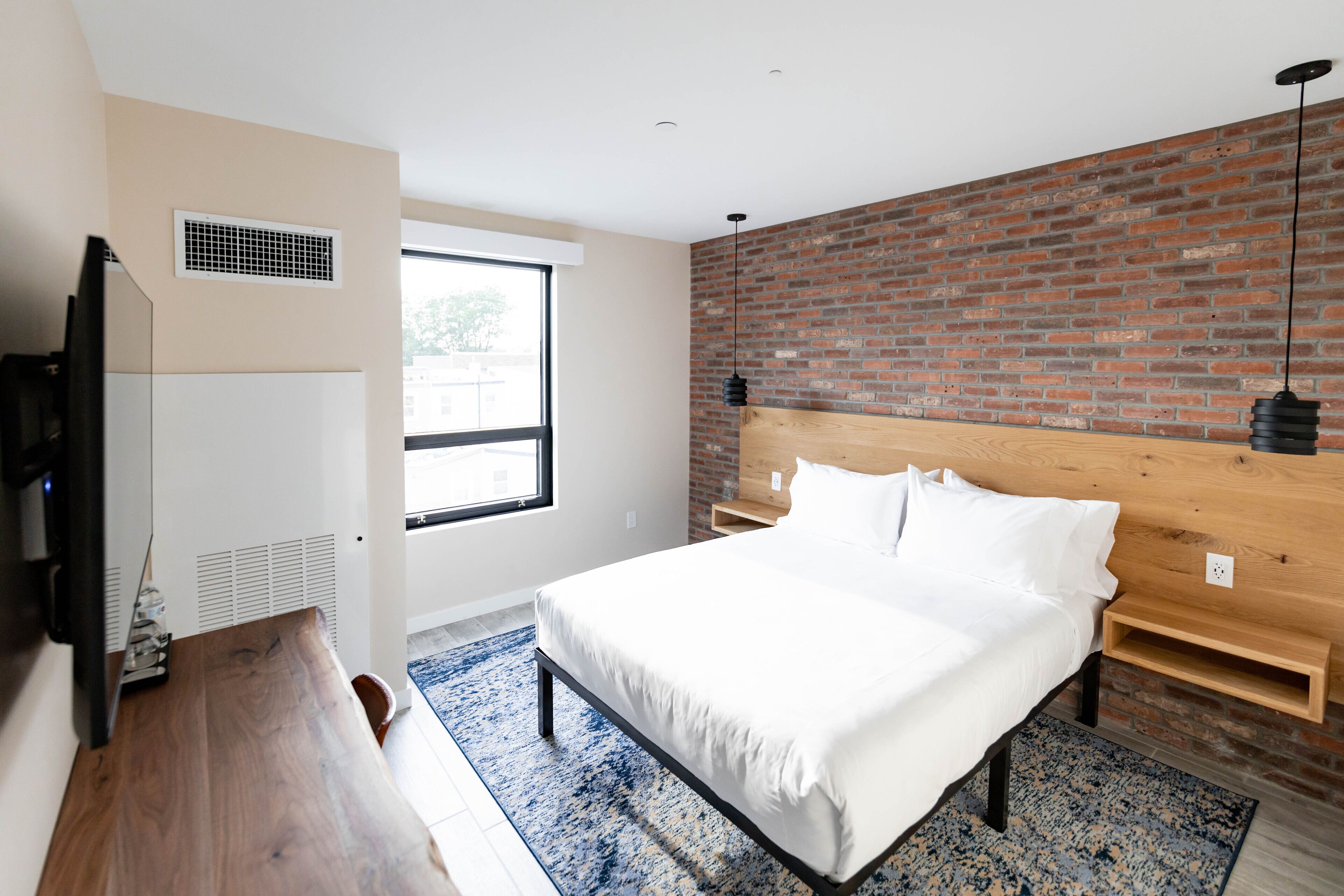 A modern bedroom with a brick wall, a wooden headboard, a large bed, a TV, and a window.