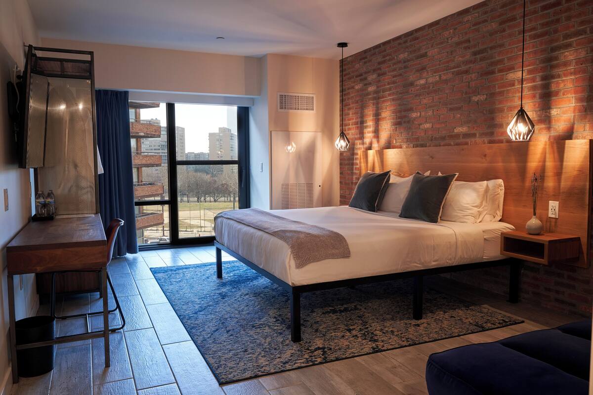 A modern bedroom with a large bed, a desk, and a view through a glass door.