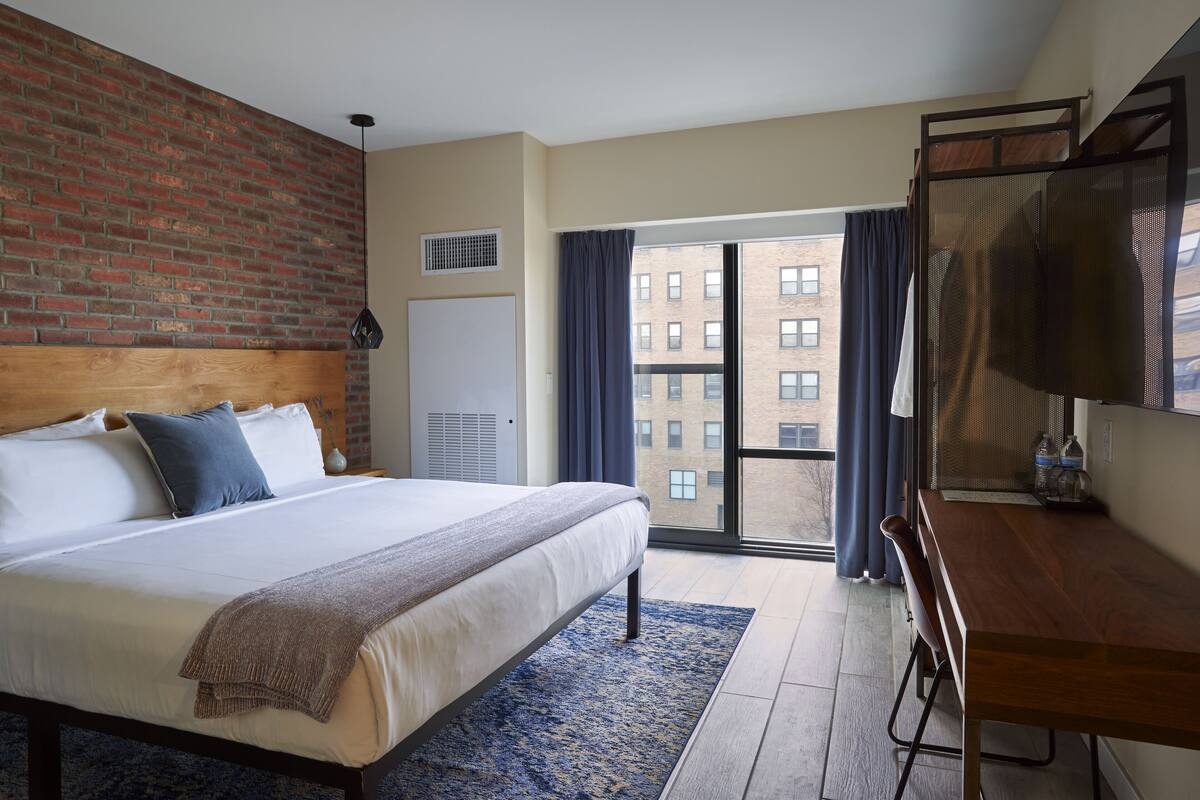 A hotel room with a large bed, a desk, a TV, a window with curtains, and a view of buildings outside.