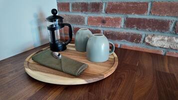 Un plateau en bois contenant une cafetière à piston, deux tasses en céramique et un torchon plié, le tout posé sur une table en bois devant un mur de briques.