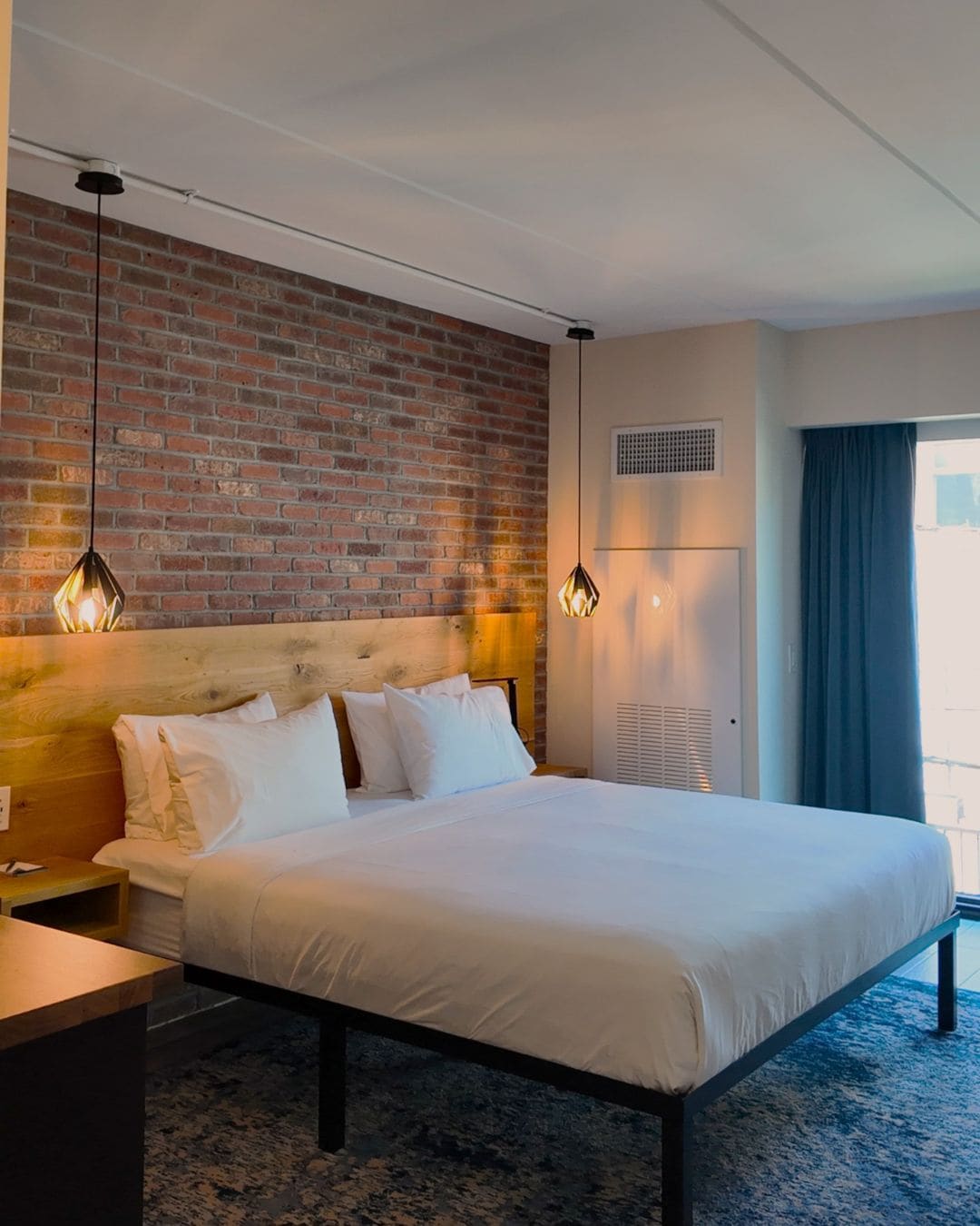 A hotel room with a brick wall, a bed with white bedding, a wooden headboard, a nightstand, and two pendant lights.