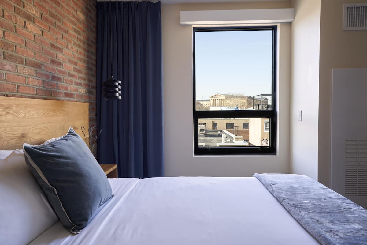 A hotel room with a large window, a brick wall, a bed with white bedding, and a blue curtain.
