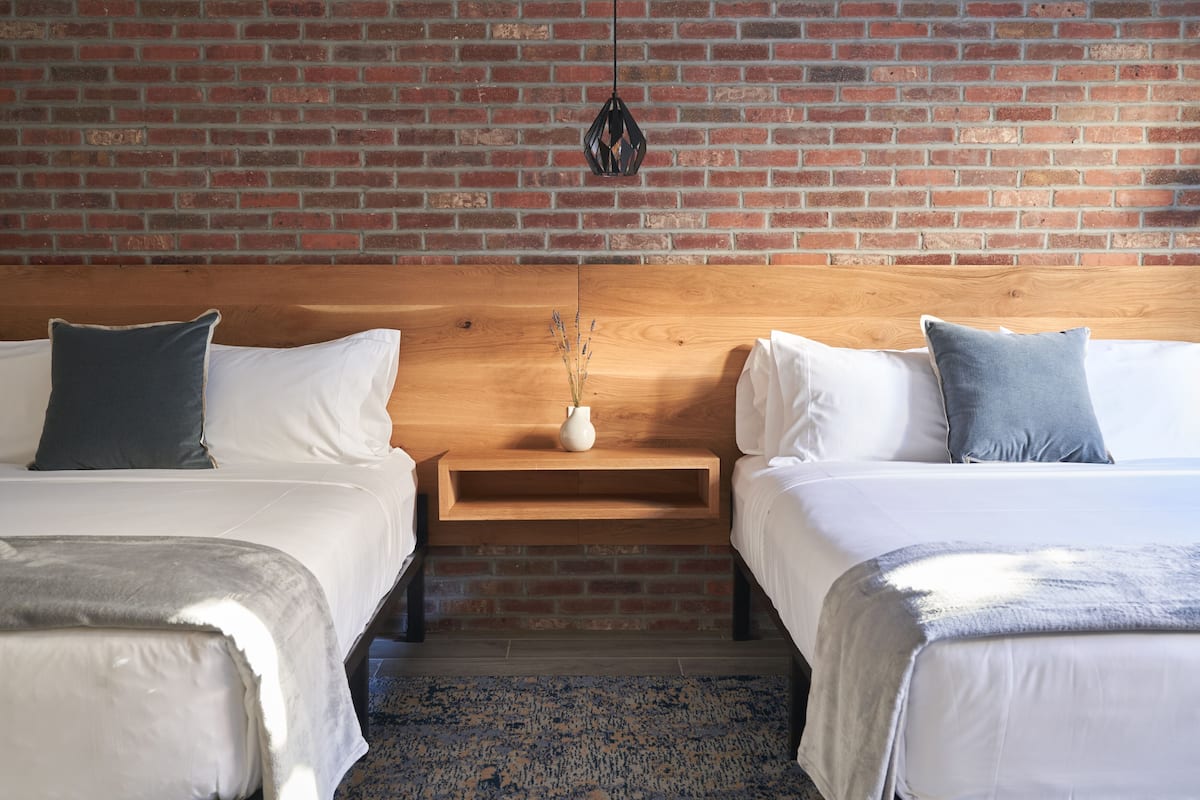 A hotel room with two beds, a wooden headboard, a brick wall, and a small nightstand with a vase.
