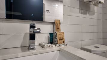 A bathroom with a white tiled wall, a chrome faucet, and a toilet. There are two brown paper bags on the counter, one labeled 'NISGUR' and the other 'FRESH MOUTH'.