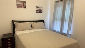 Un lit bien fait, recouvert d’une courtepointe matelassée, une table de chevet en bois avec des tiroirs, et deux tableaux encadrés accrochés au mur.