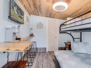 A compact room with a bunk bed, a wooden table with stools, a small desk, and a ceiling fan.
