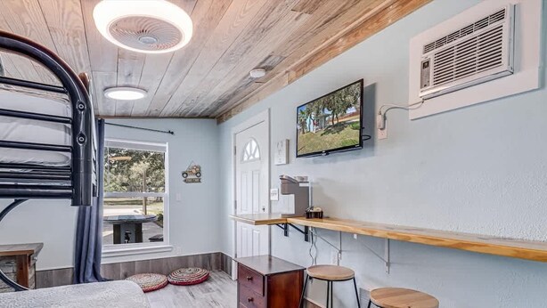 A room with a bed, a TV mounted on the wall, a wooden desk, and a bar area with stools. - G & J Paradise (Rockport)