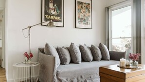 Een moderne woonkamer met een grijze bank, een rond bijzettafeltje met een lamp, en kunstwerken in lijsten aan de muur.