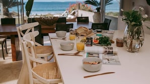 A dining table with breakfast items, a view of the ocean, and a tropical plant. - 'Atra' - Jervis Bay Beach Front Holiday Home (Callala Beach)