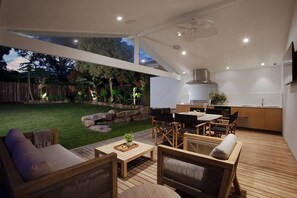 Eine moderne Küche mit eingebautem Herd und Dunstabzugshaube, ein Essbereich mit Holzmöbeln sowie eine Terrasse mit Holzboden und Gartenmöbeln.