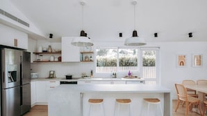A modern kitchen with white cabinetry, stainless steel appliances, a central island with bar stools, pendant lighting, and a view of the outdoors through large windows.