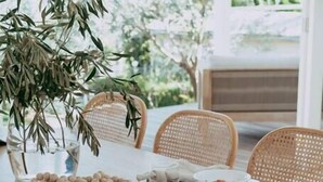 A dining table set with various dishes, including bread, fruits, and vegetables, in a bright, naturally lit room with wicker chairs.