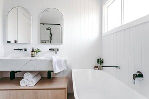 Ein modernes Badezimmer mit einem weißen Waschtisch, zwei Spiegeln, einer Badewanne und einem Fenster.