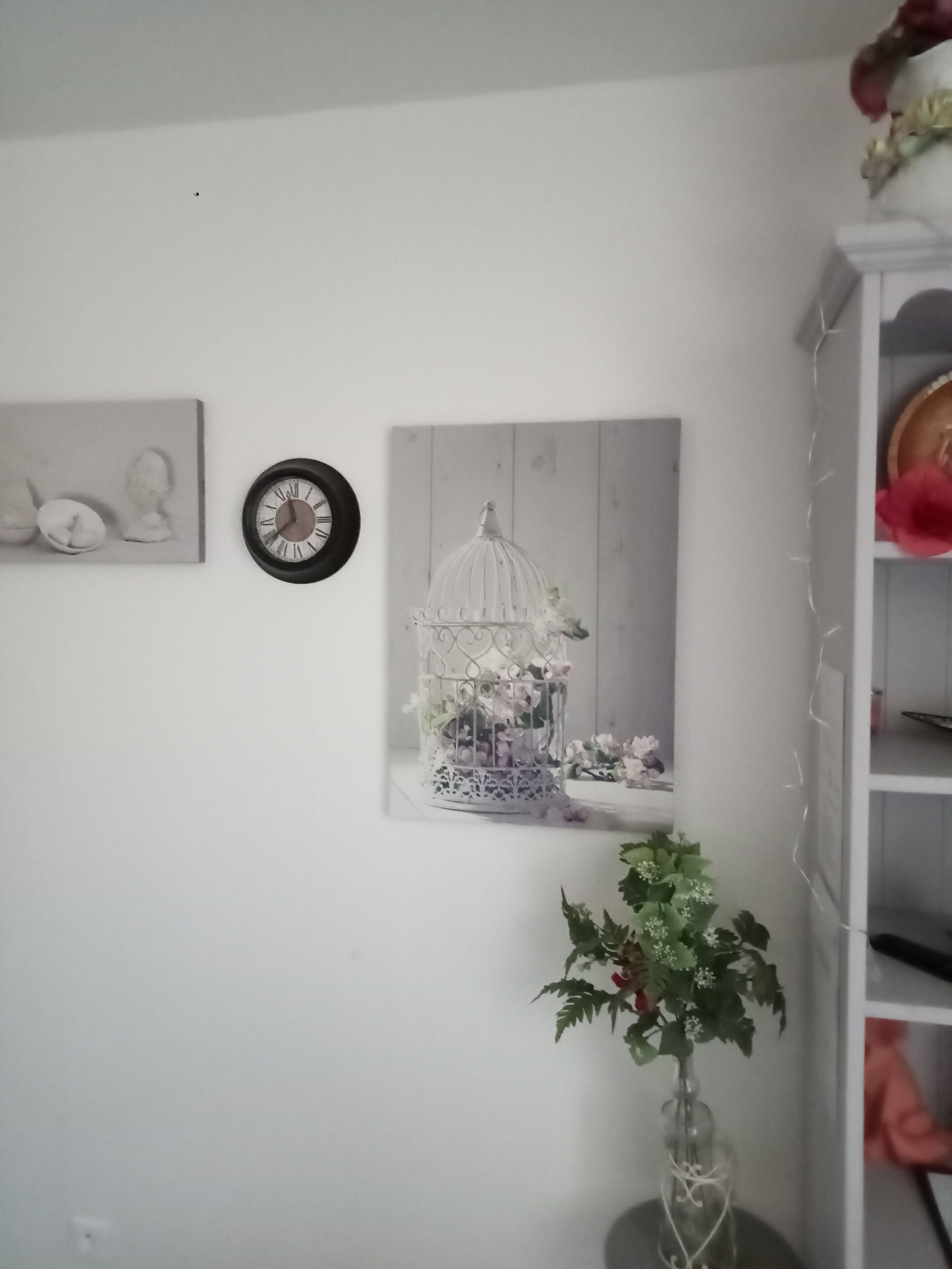 Una habitación con pared blanca, un reloj, un cuadro enmarcado, una jaula decorativa y una planta en maceta.