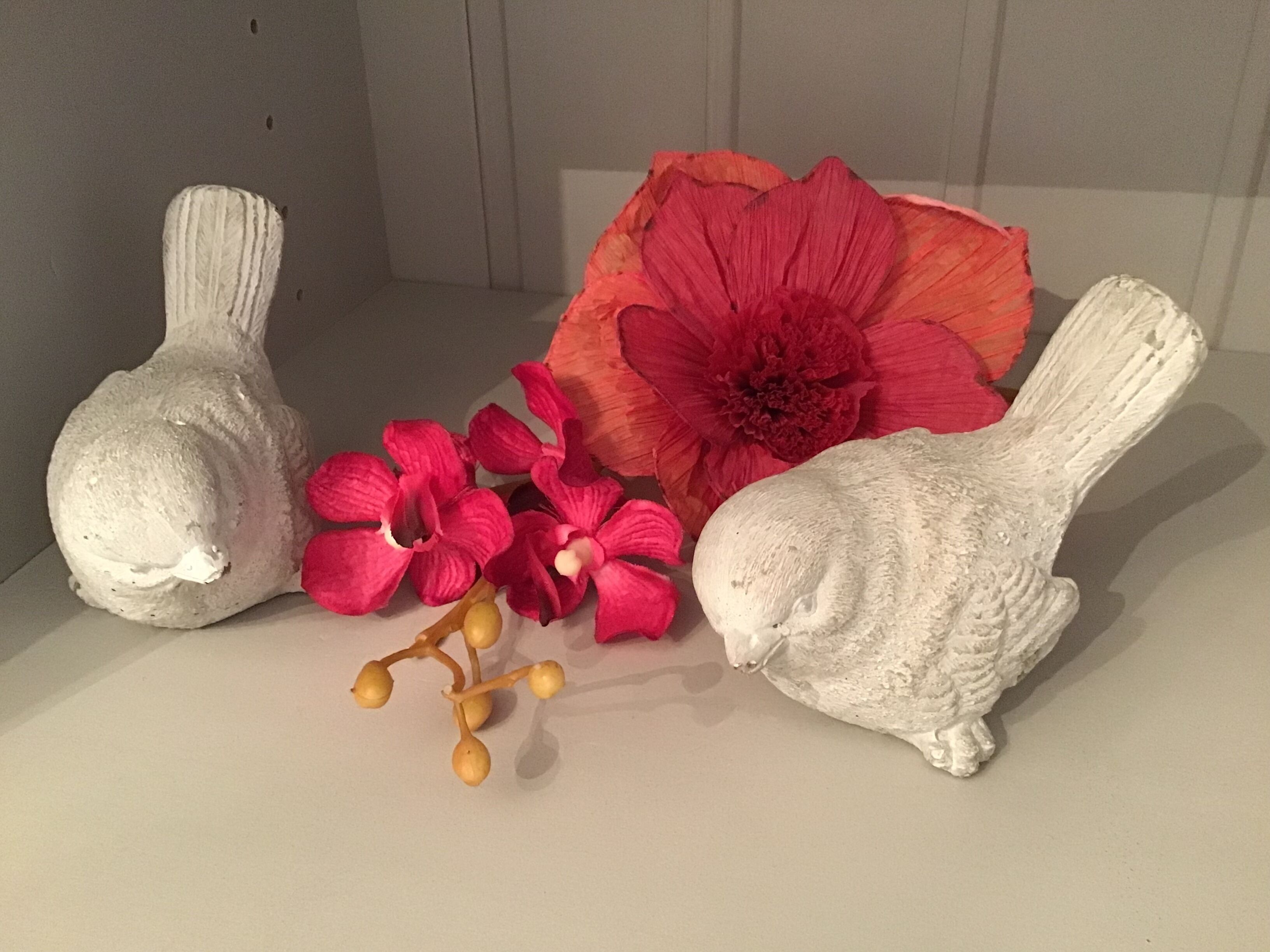 Three white ceramic sculptures, one of which is a rabbit, placed on a flat surface with a large red flower and smaller pink flowers in the foreground.