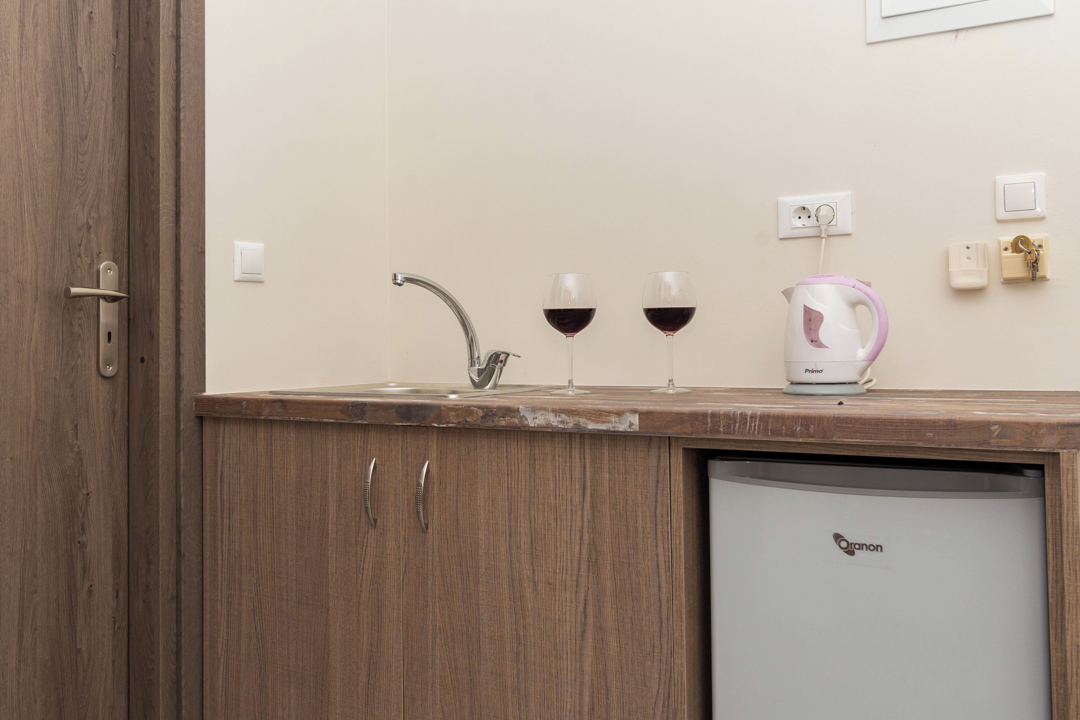 A kitchen with a sink, two wine glasses, a kettle, and a refrigerator.