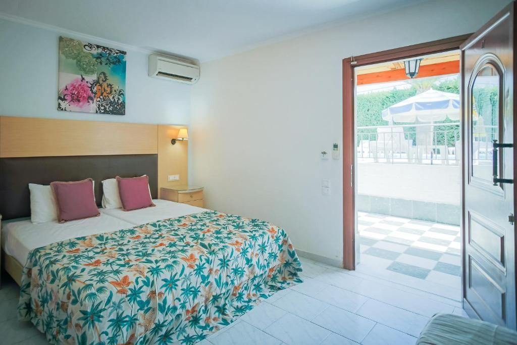 A hotel room with a bed, a nightstand, an air conditioning unit, a painting on the wall, and a balcony with a checkered floor.