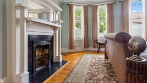 A room with a fireplace, wooden flooring, a patterned rug, and a view through a window with curtains.