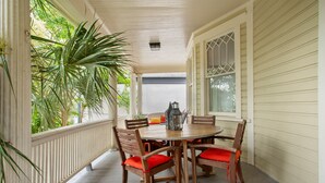 A covered porch with a wooden table and chairs, surrounded by white railings and a large potted plant.