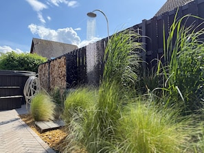 A modern outdoor shower area with a stone wall, surrounded by tall grasses and a wooden deck.