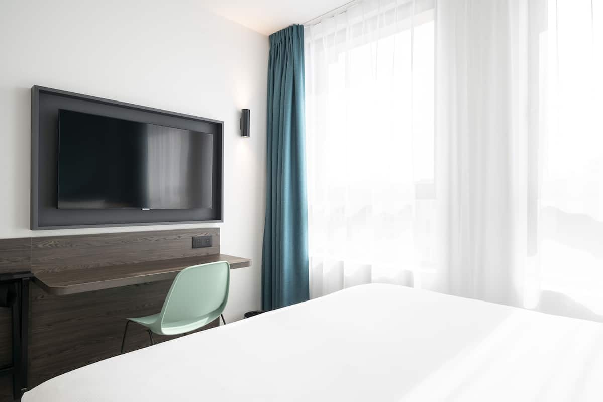 Habitación de hotel con cama, televisor montado en la pared, escritorio con silla y ventana con cortinas.