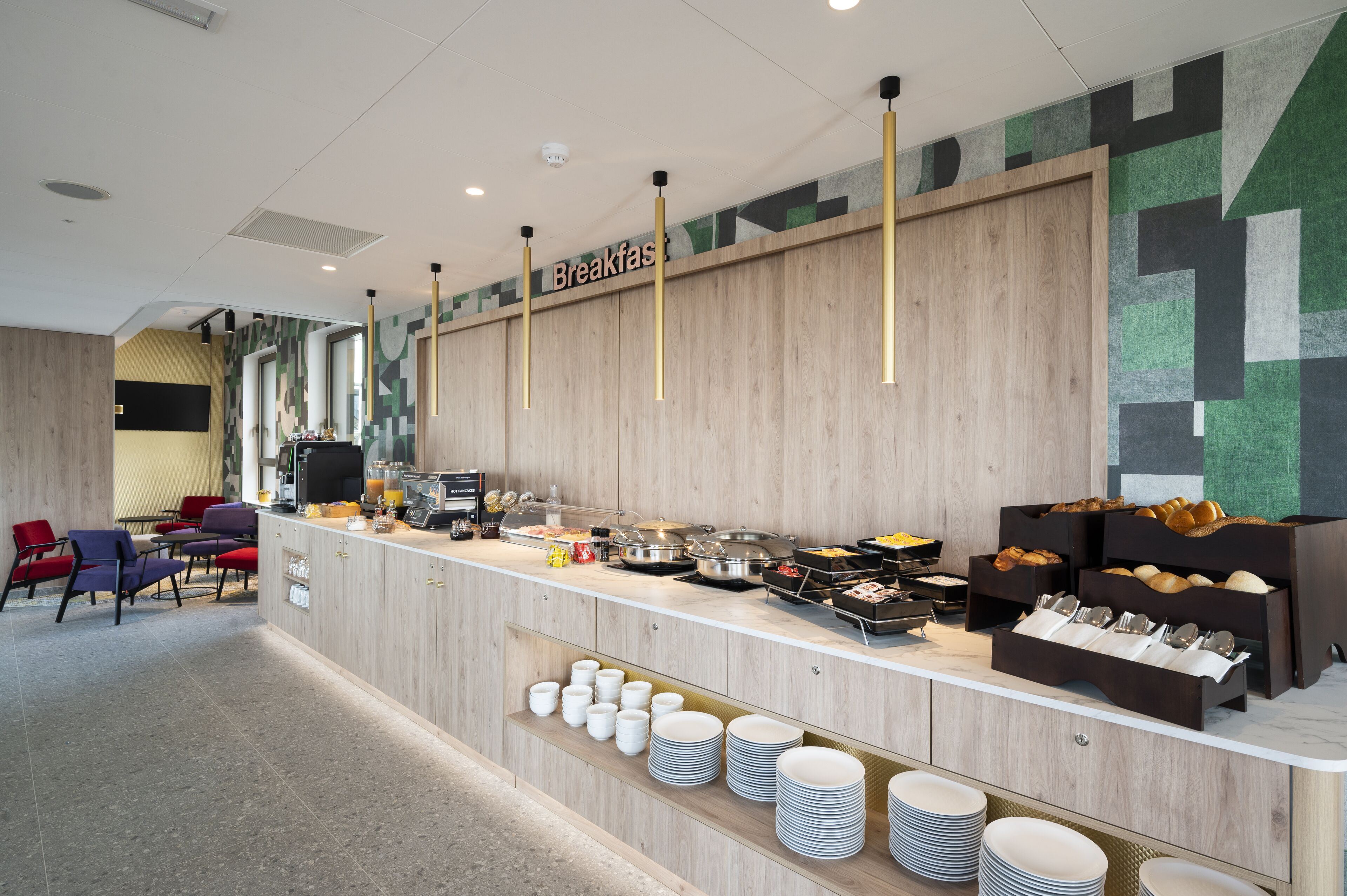 Ein Frühstücksbuffet mit verschiedenen Speisen und Besteck, eine Sitzecke mit Stühlen und eine Wand mit geometrischen Mustern.