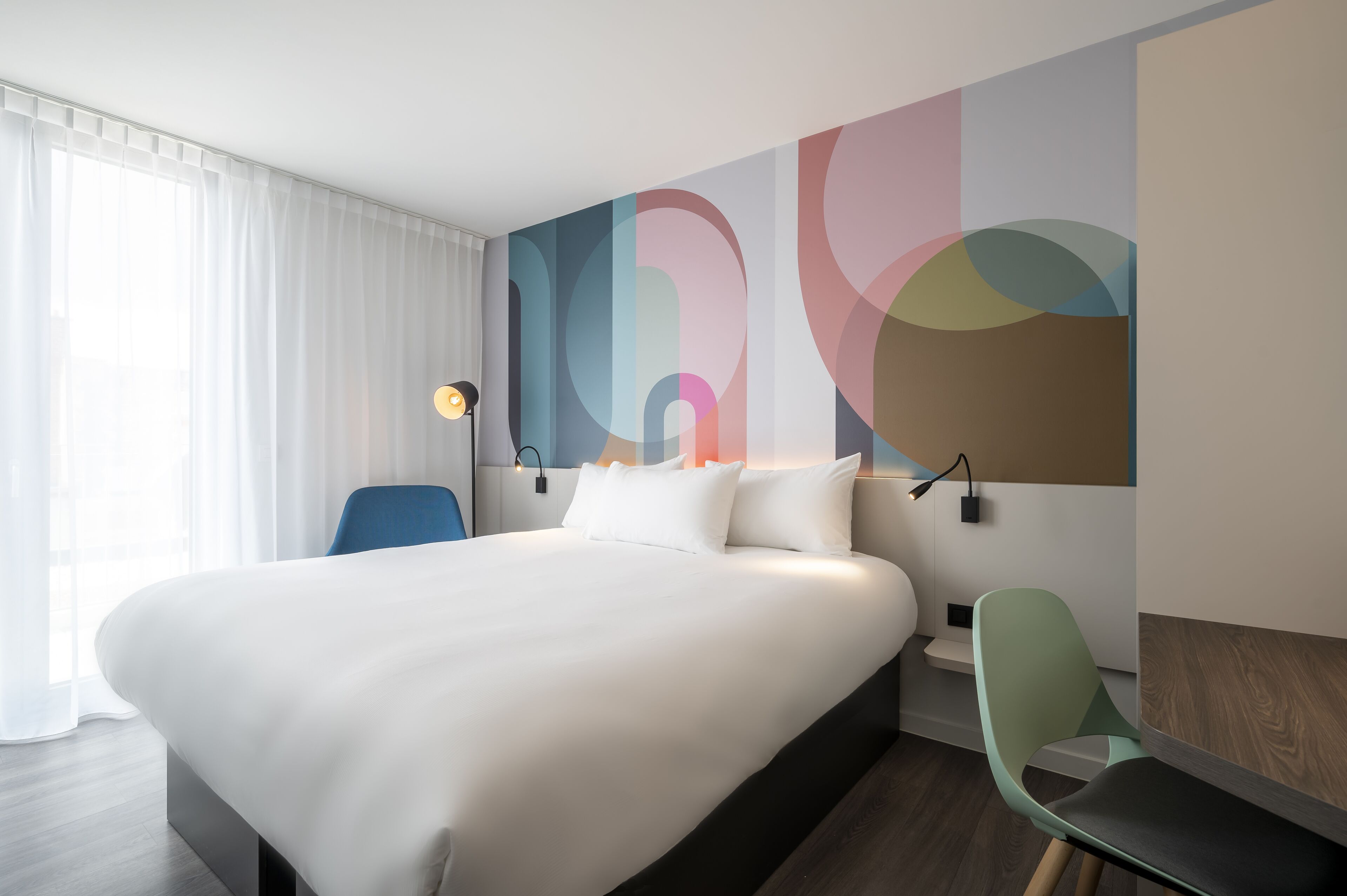 Una habitación de hotel moderna con una cama grande, un escritorio y una silla. En la pared, un cuadro abstracto de gran tamaño y una ventana con cortinas blancas.