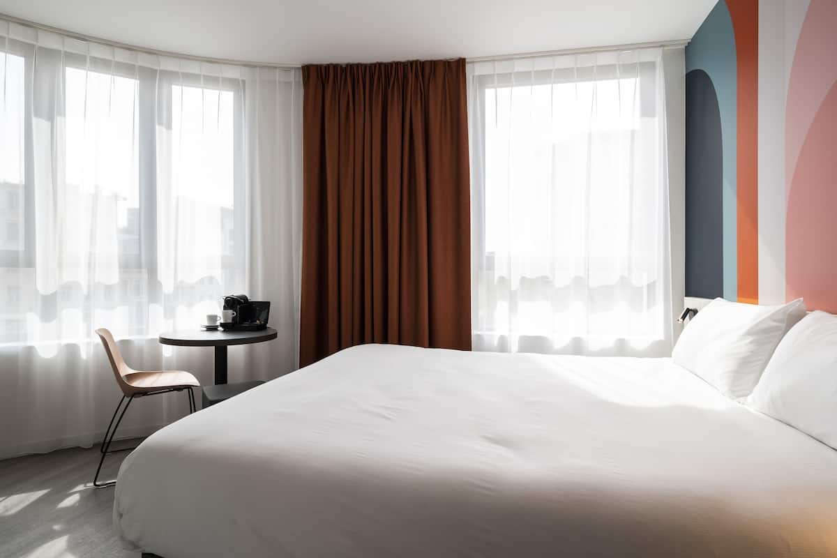 Habitación de hotel con una cama grande, una silla, una mesita con cafetera y vistas a la ciudad a través de cortinas translúcidas.