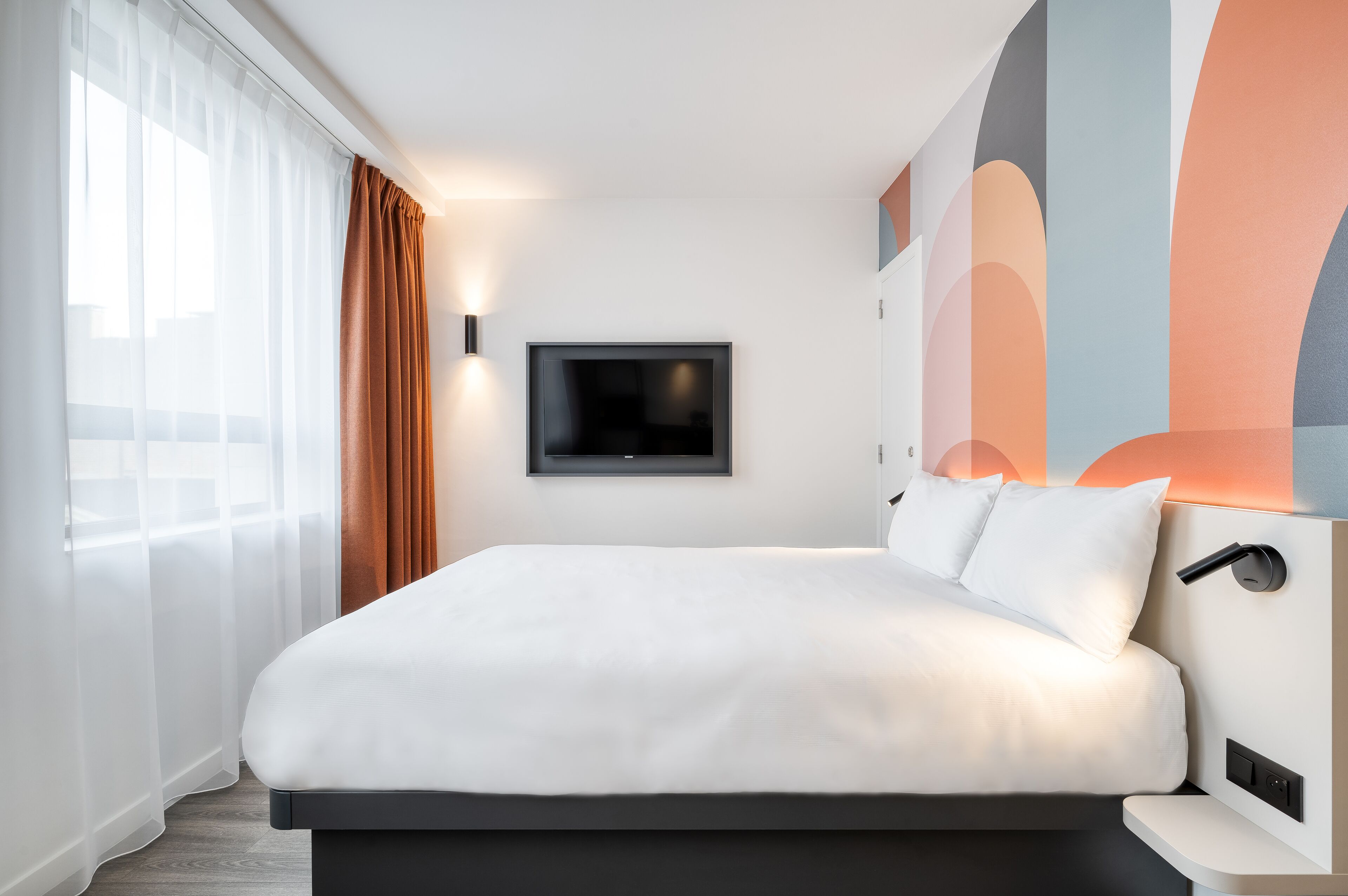 Une chambre d’hôtel moderne dotée d’un grand lit, d’un téléviseur à écran plat fixé au mur et d’une grande fenêtre avec des rideaux légers.