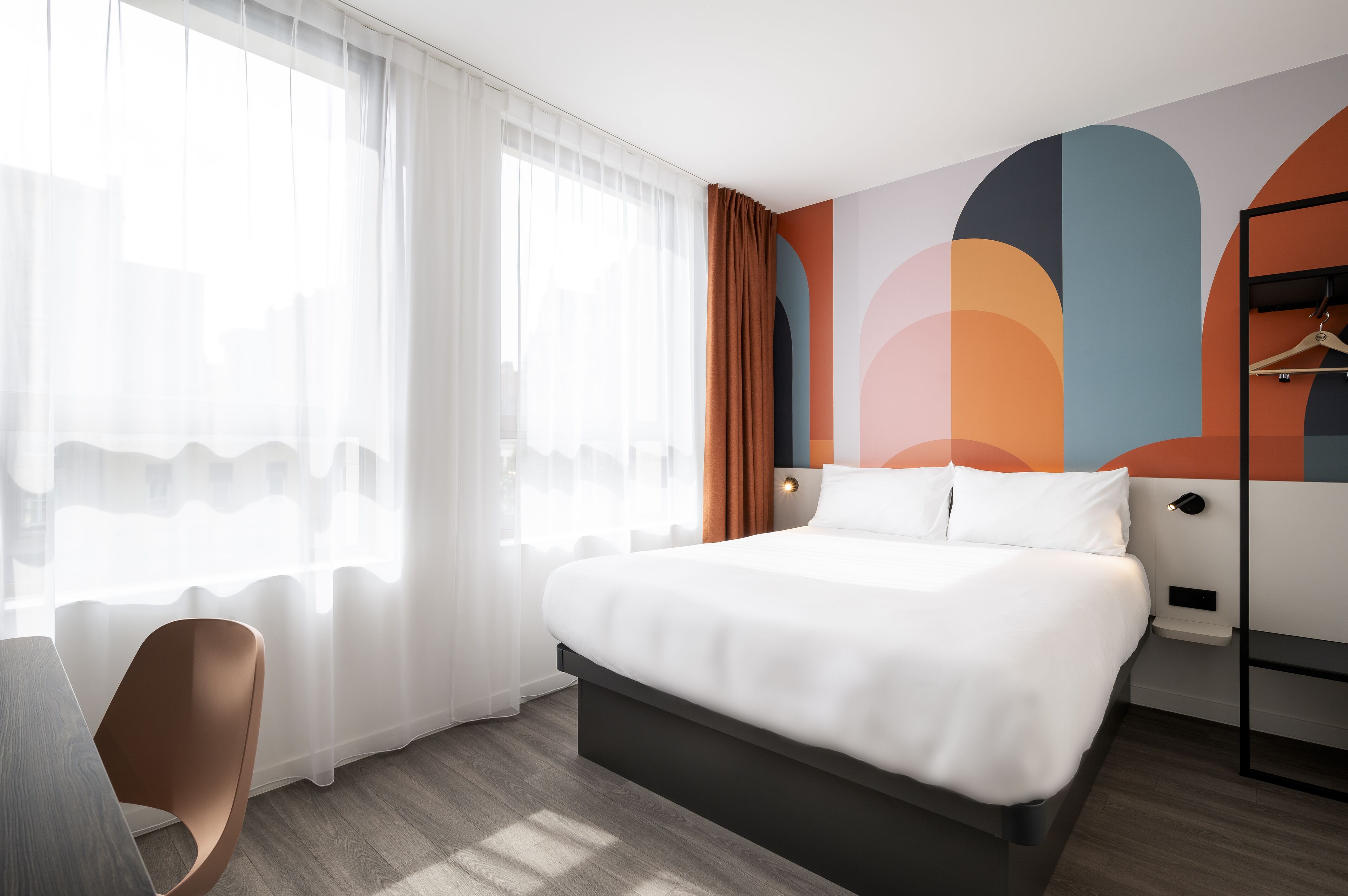 A modern hotel room with a large bed, a chair, a wardrobe, and a wall with abstract art.