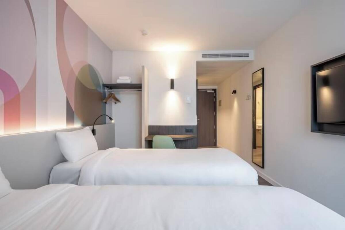 Habitación de hotel con dos camas, un escritorio, una silla, un televisor y una pared con arte abstracto.